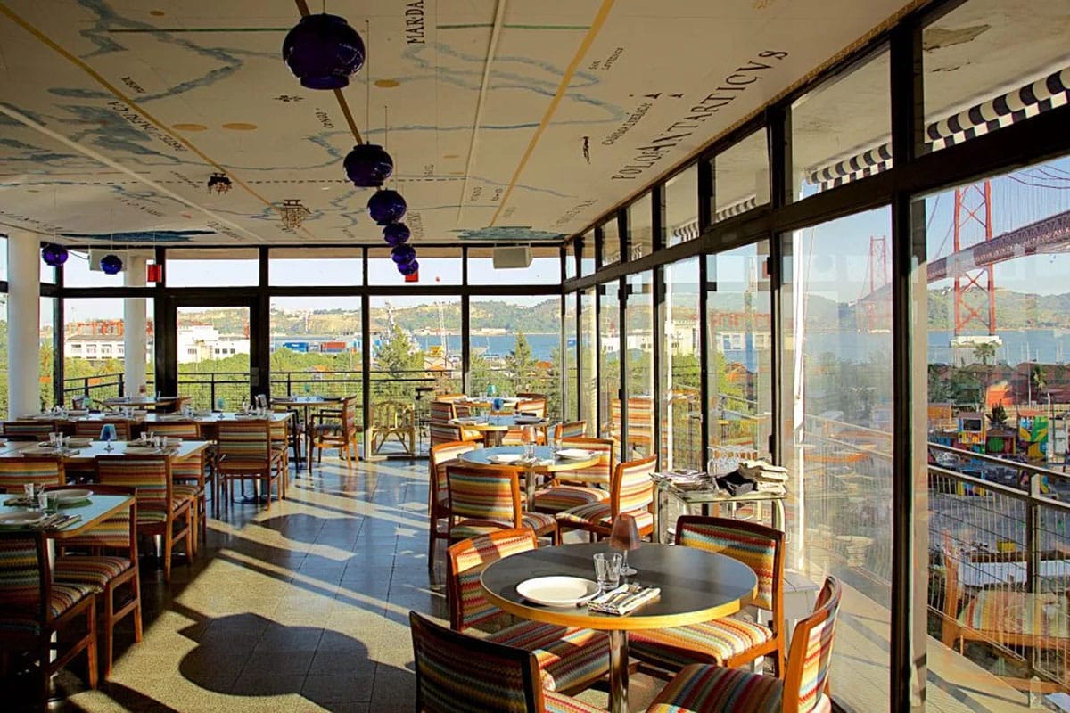 colorfully upholstered wooden chairs at tables in a glass-walled room overlooking Lisbon's river, city, and bridge.
