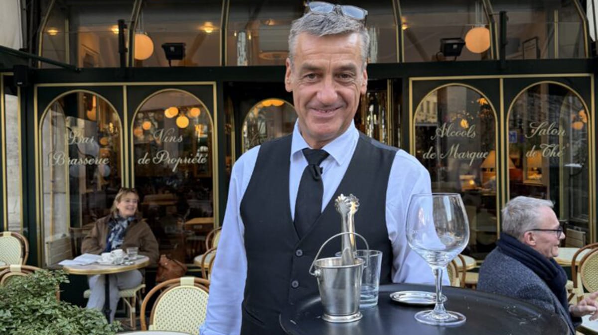French waiter dressed in a vest and tie holding a platter with a wine glass.