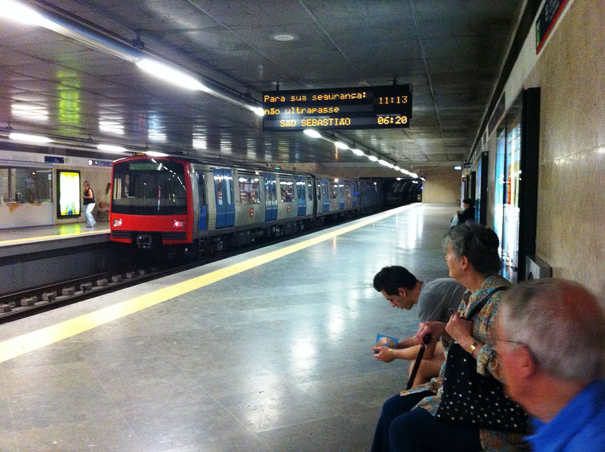 Lisbon metro train approaching the station with travelers seated on a bench nearby.