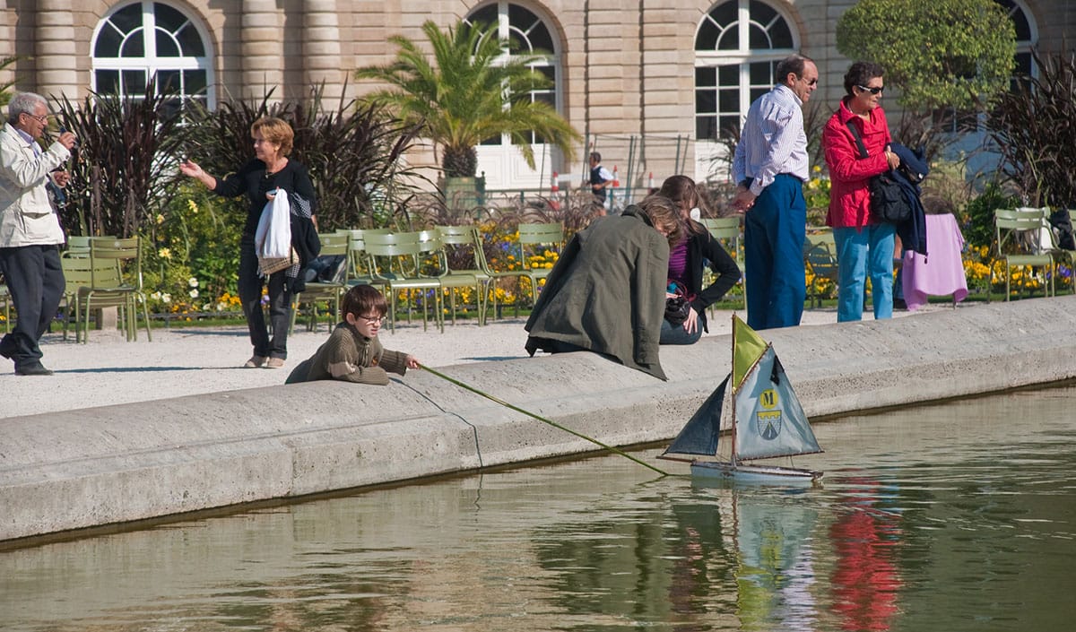 boy pushing a sailboat on a pond with a long stick while adults look on.