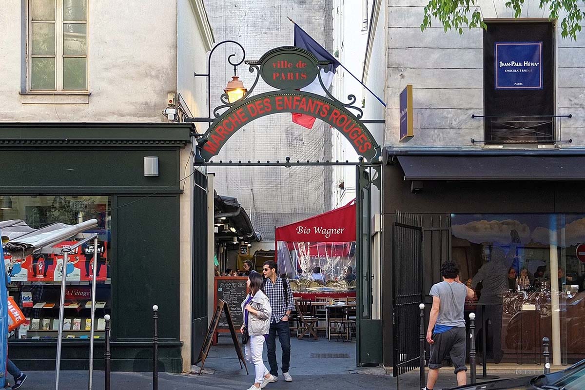 people walking outdoors under the arched entryway to a French food market.