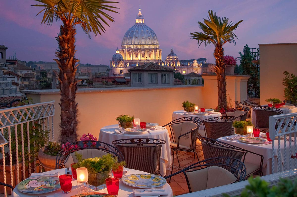 candlelit, tablecloth-covered tables on a rooftop with palm trees overlooking a cathedral dome.