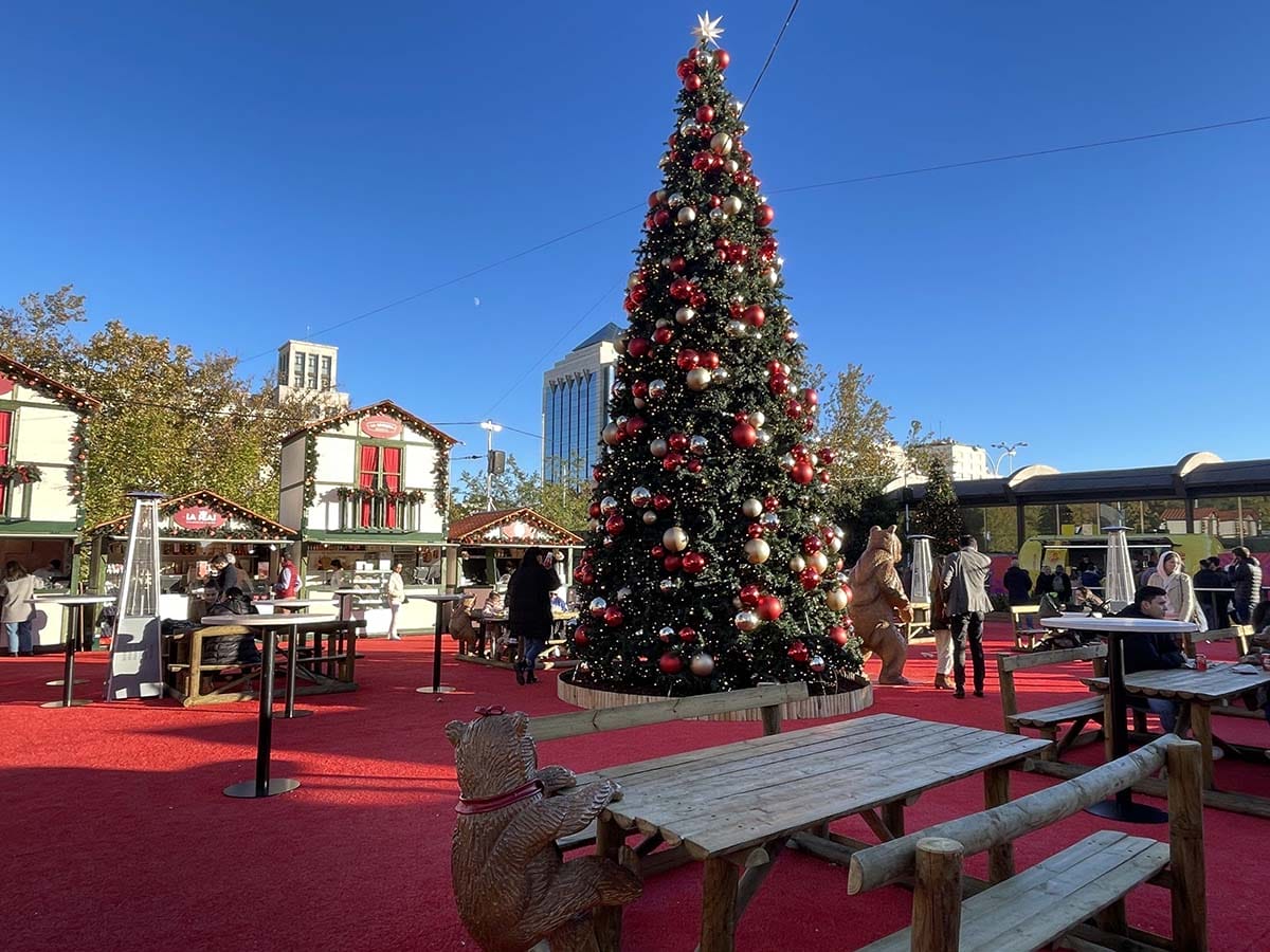 large Christmas tree in the center of a market with booths and tables around it.