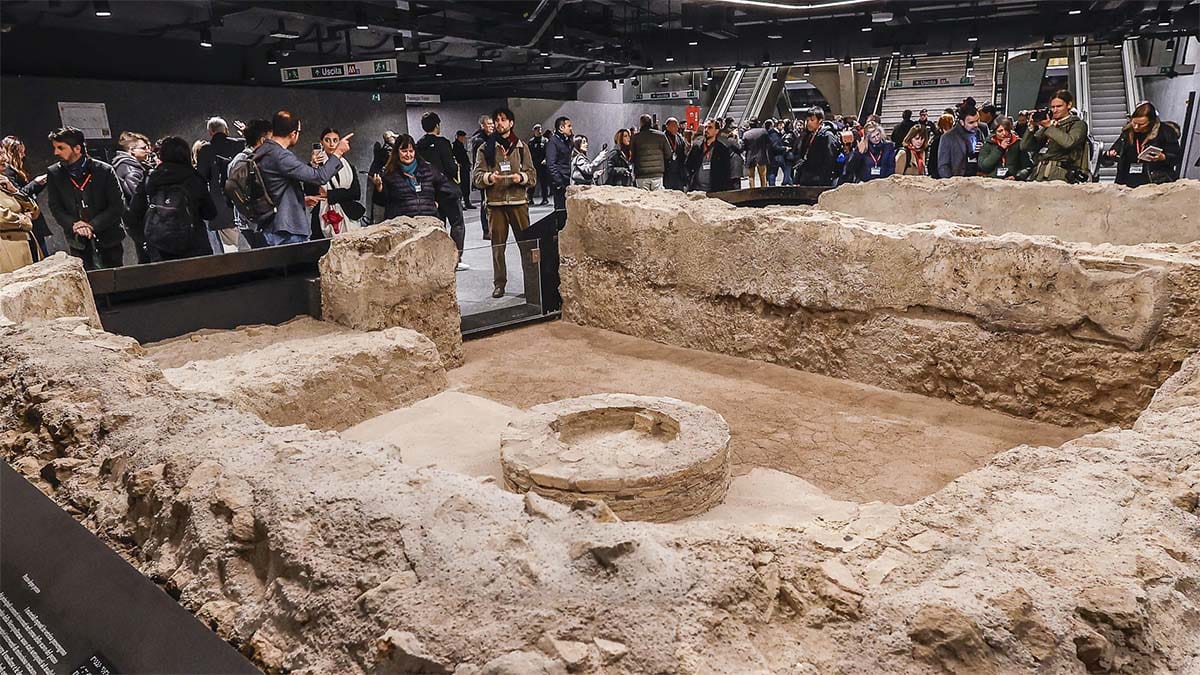 group of journalists and metro passengers gathered around ancient Roman ruins in a subway station.
