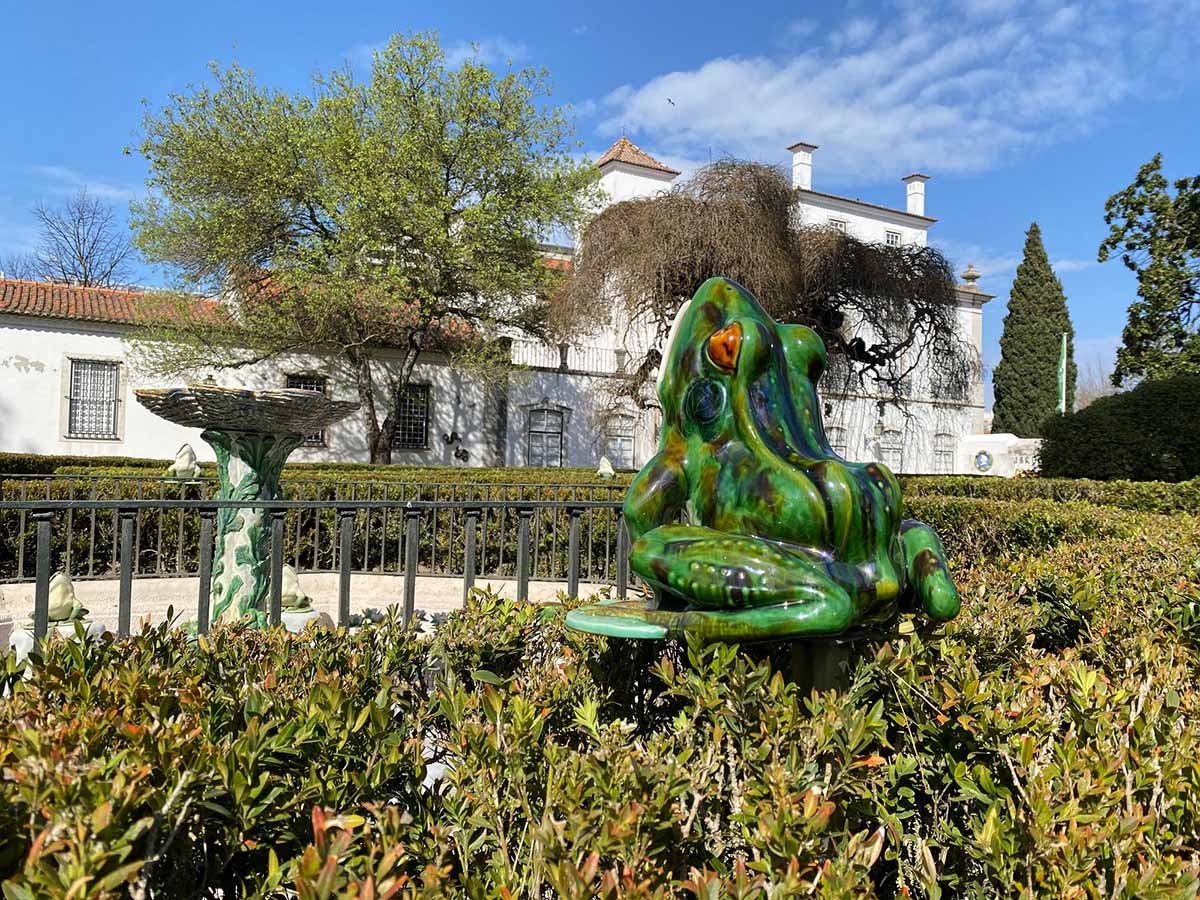 large ceramic frog on a pedestal in a hedge facing a fountain.