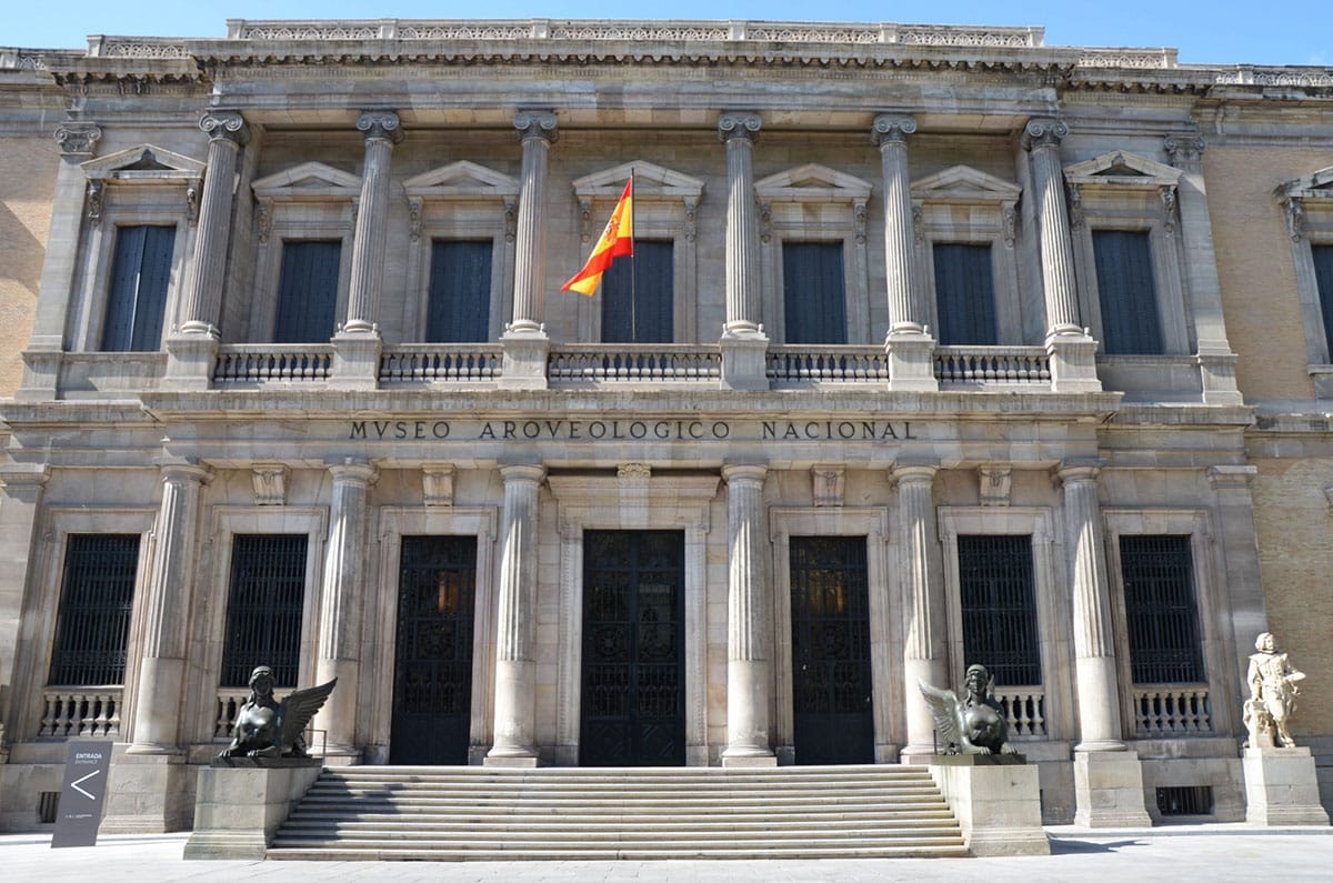 large stone building with columns, a balcony, and statues.