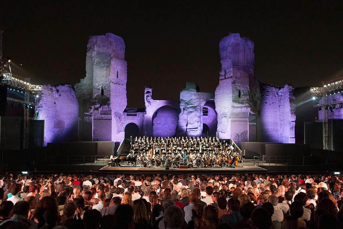 symphony on stage with illuminated Roman ruins in the background and a large audience outside.