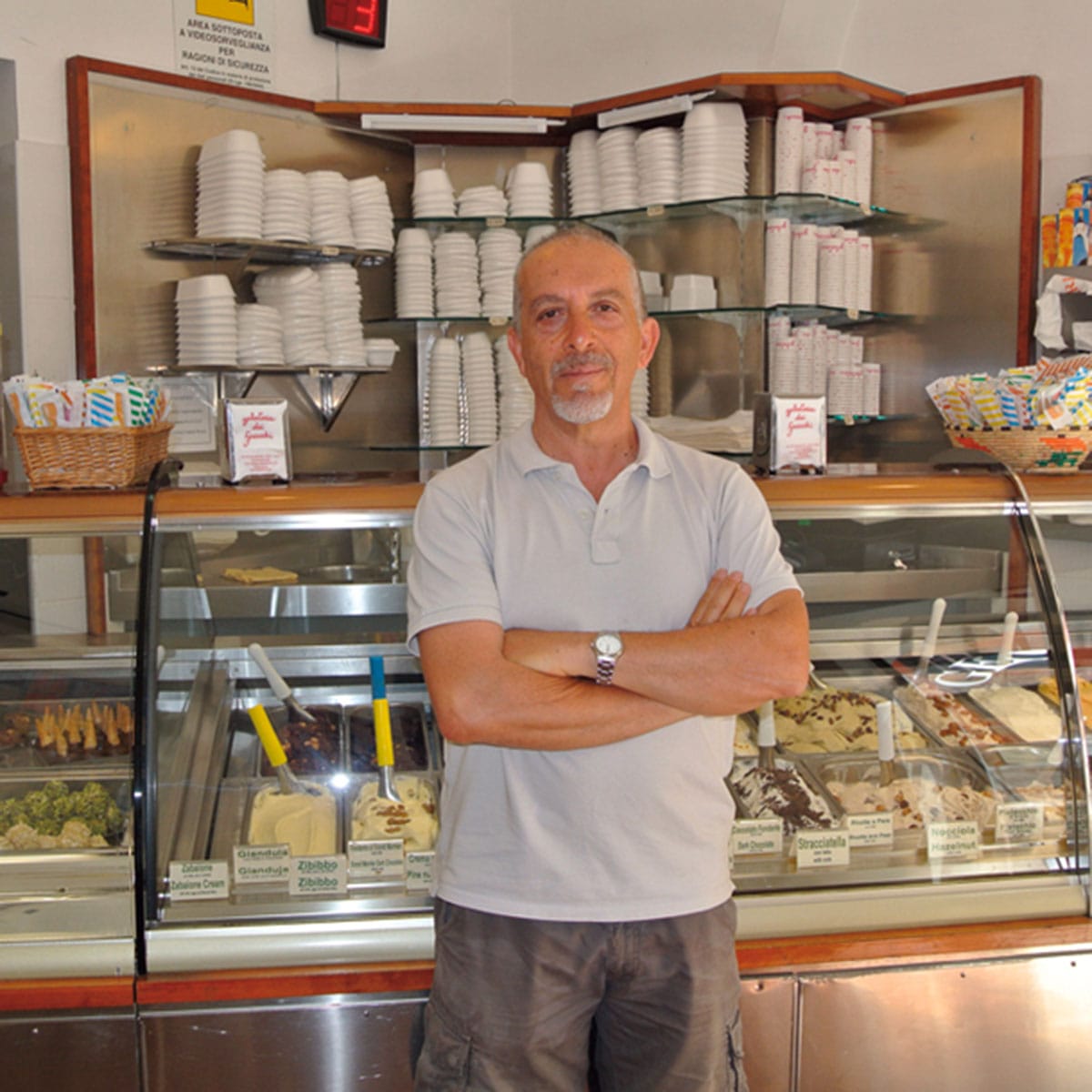 man posing in front of a glass case of gelato in serving containers.