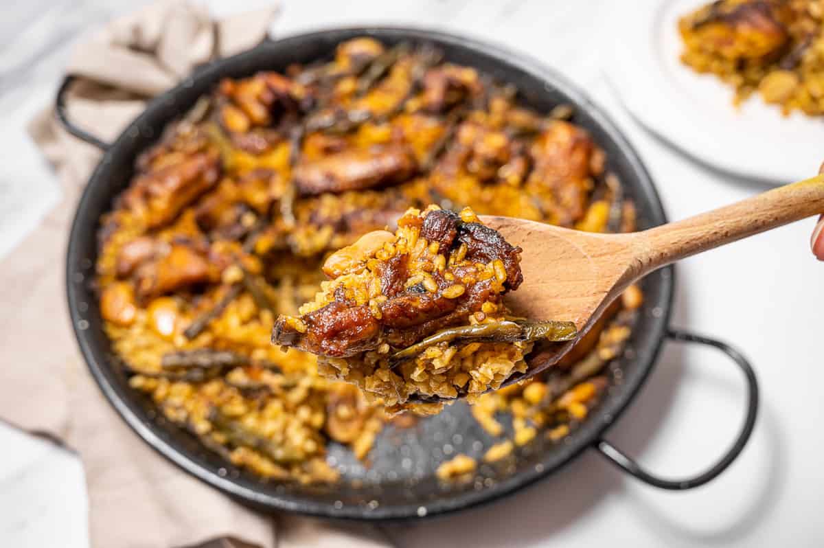 wooden spoon of paella valenciana with the pan in the background on a marble table.