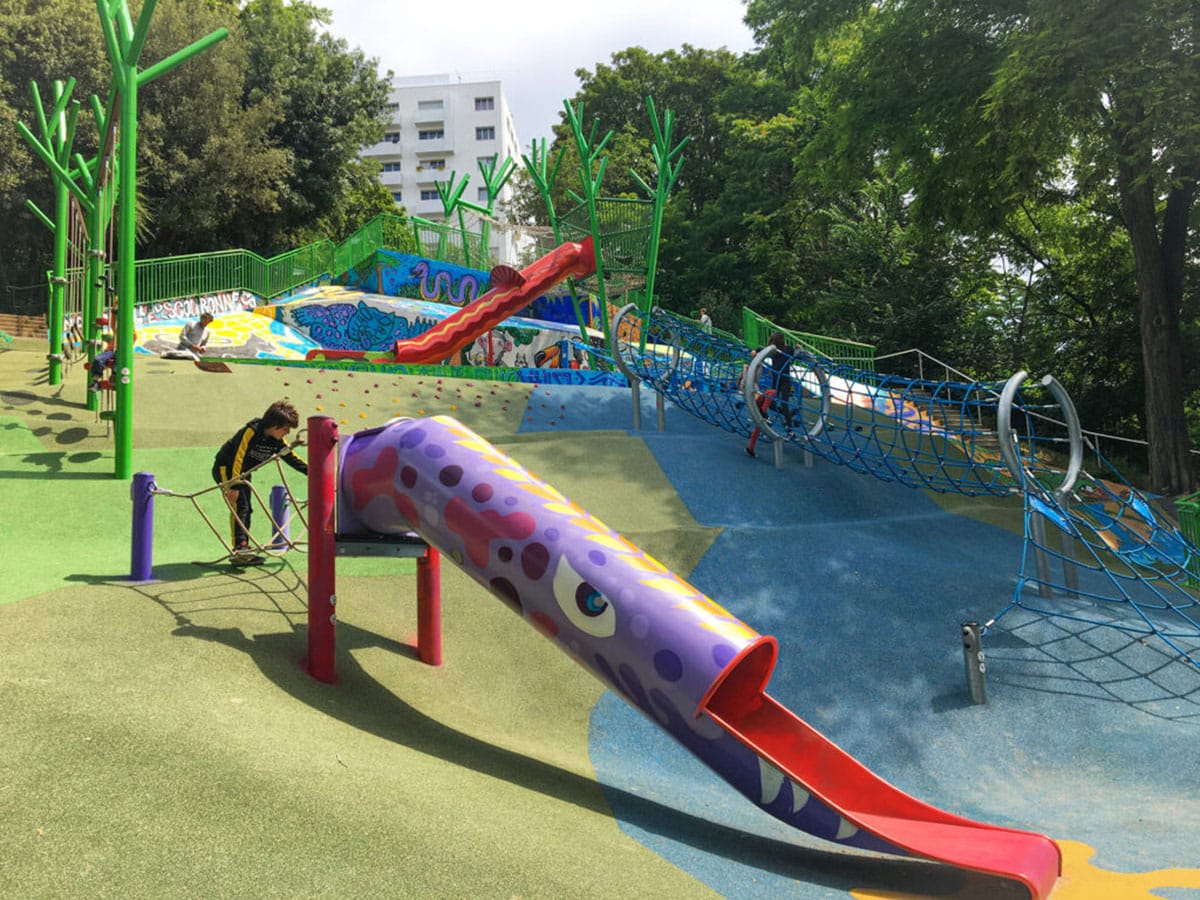 colorful slides and playground equipment on a hill surrounded by trees.