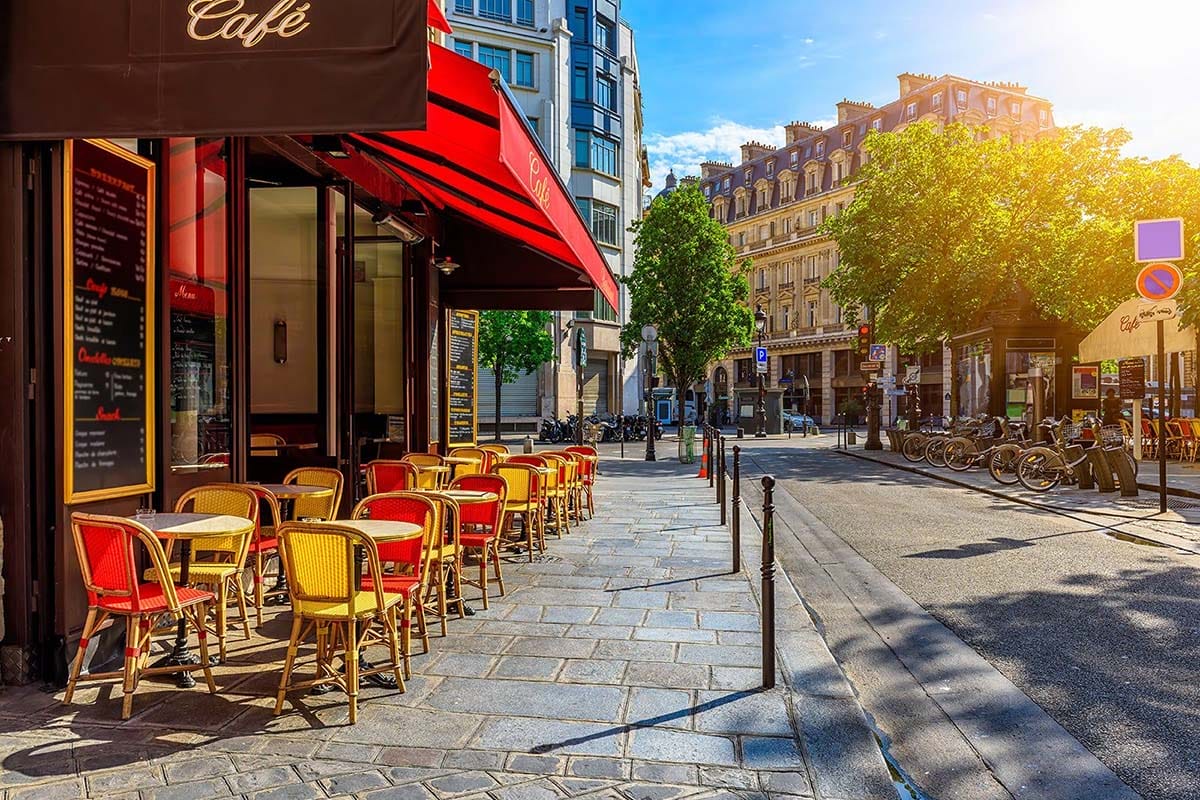 paved street lined with trees and tall buildings with a restaurant on the sidewalks.