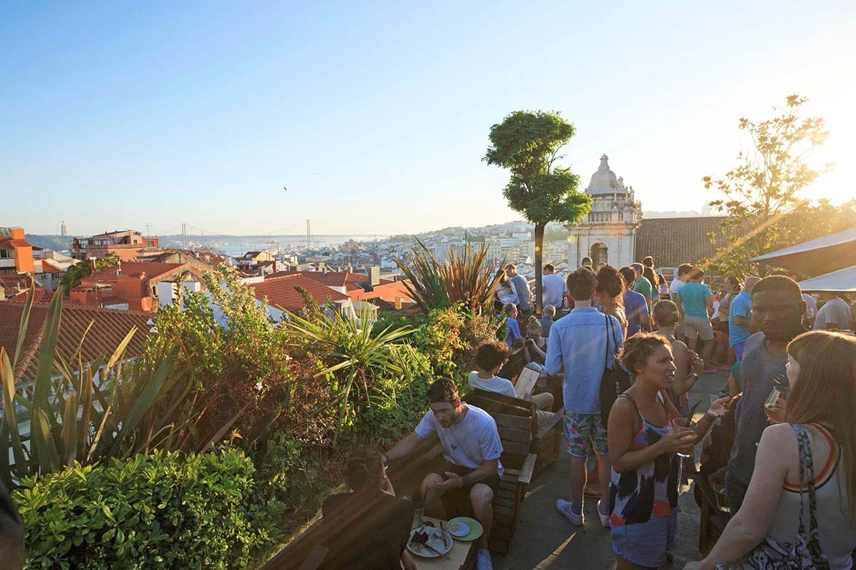 people at a rooftop bar near bushes and trees overlooking Lisbon.