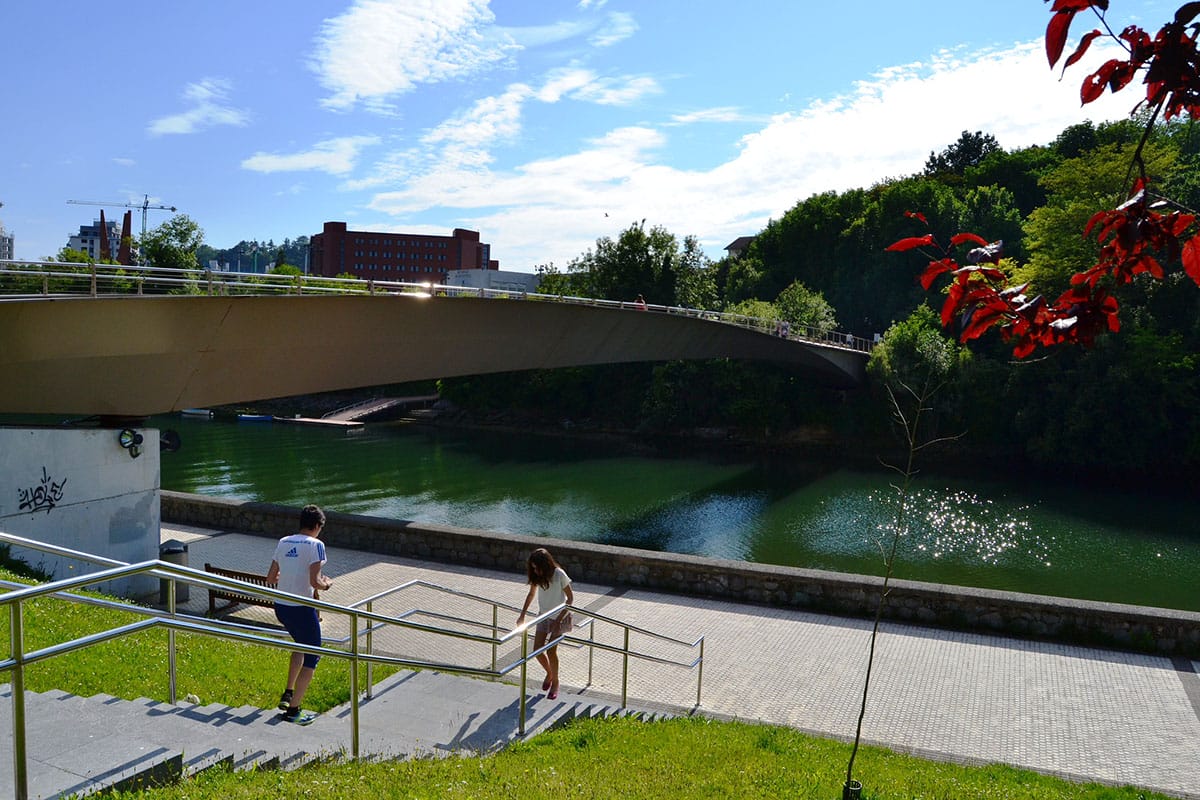 paved walkway beside a tree-lined river with a bridge over it.