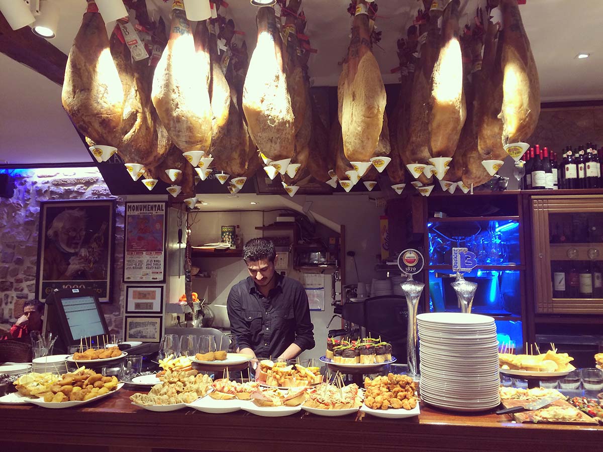 server preparing several platters of pintxos at a wooden bar underneath racks of large hams hanging from the ceiling.
