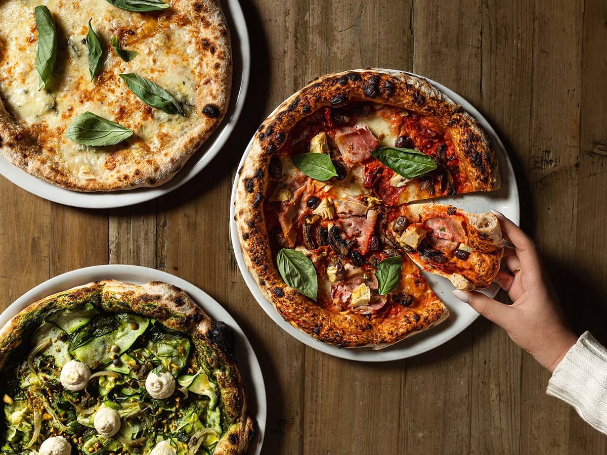 person taking a slice of pizza from a plate with two other pizzas on a wooden table.