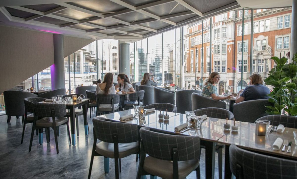 people eating at small tables surrounded by gray upholstered chairs with a great view of the street below.
