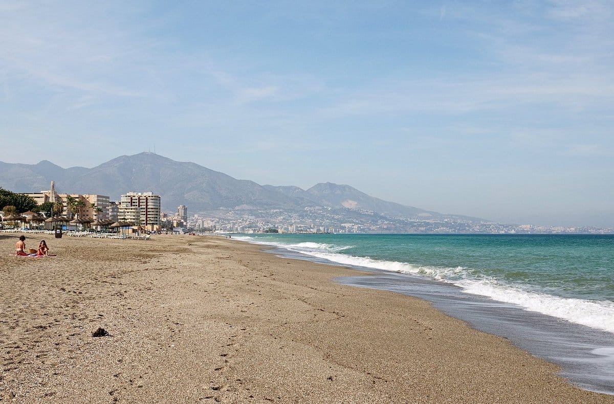 brown sand beach with blue water and mountains and a city in the background.