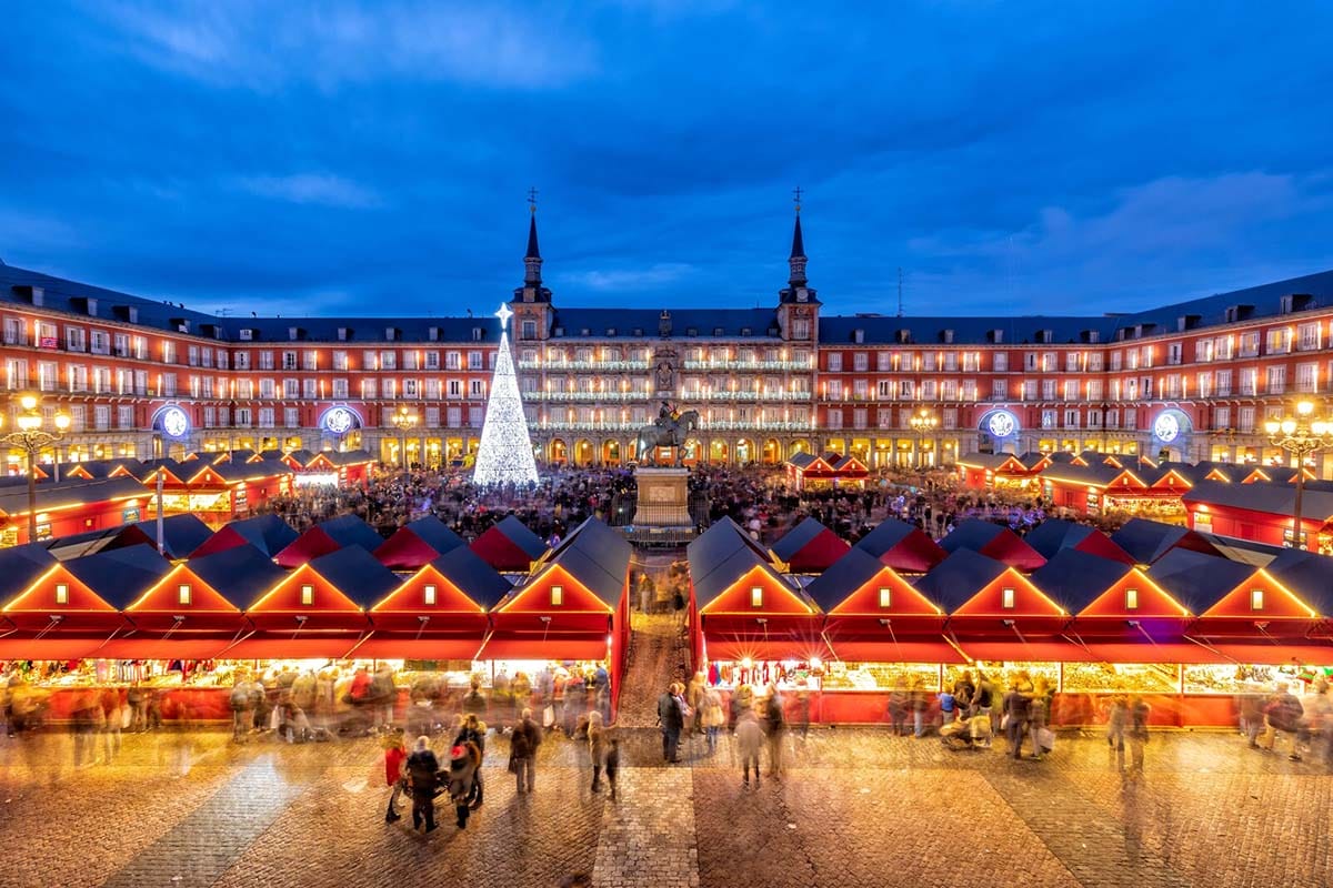 a busy plaza filled with illuminated market stalls and a lit Christmas tree.