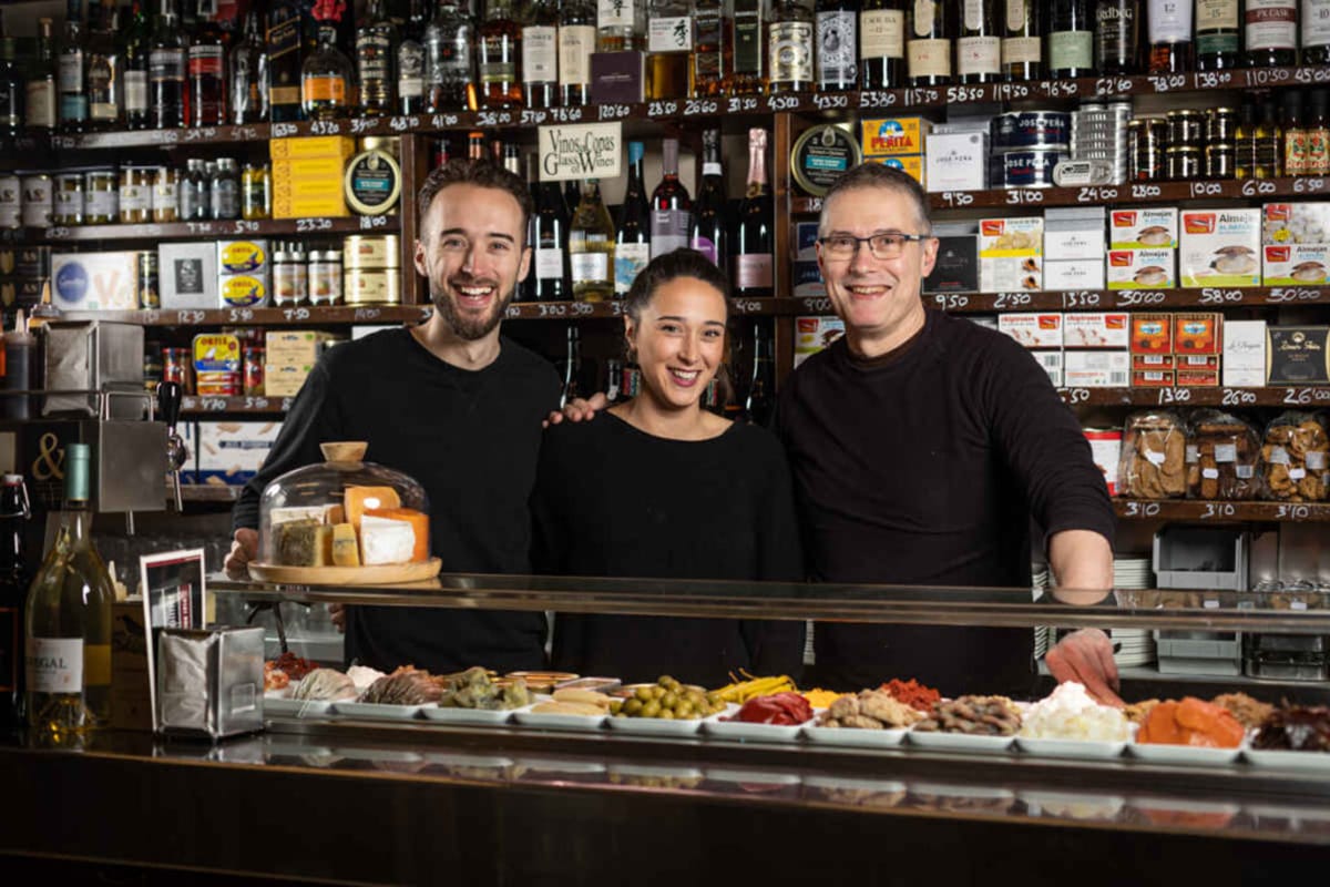 three people in front of a shelf of food and drinks with a tapas spread in front of them.