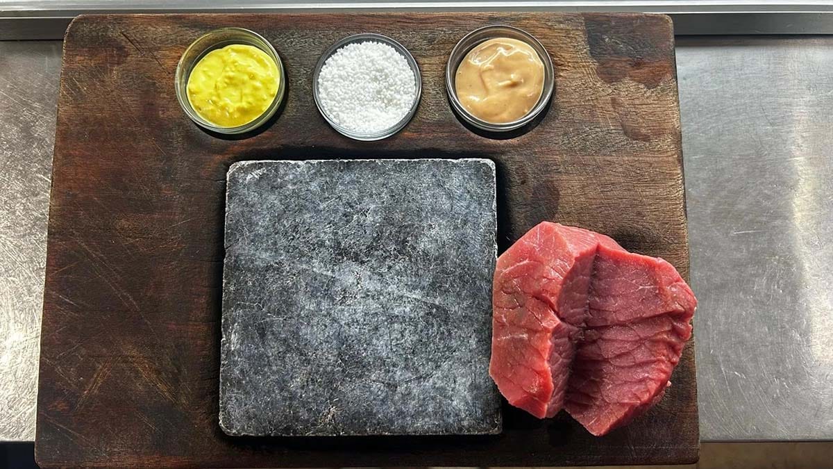 piece of sliced beef on a wooden board with a hot stone in the center, two bowls of sauces, and one bowl of salt.