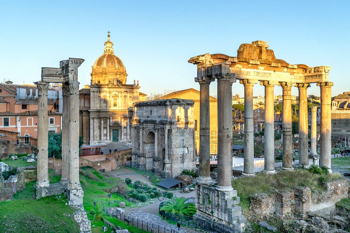 ruined Roman columns with an arch in the center and a historic church seen at sunset.