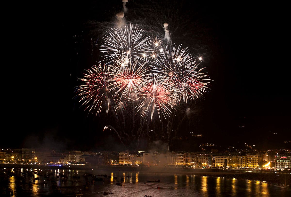 five large white and red fireworks exploding over a beach near a city at night.