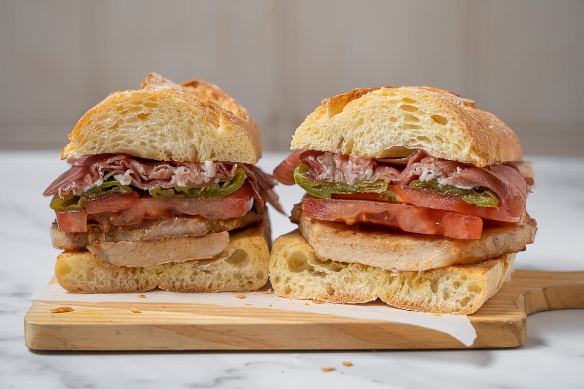large ham and pork sandwich with tomato and peppers cut in half on a cutting board.