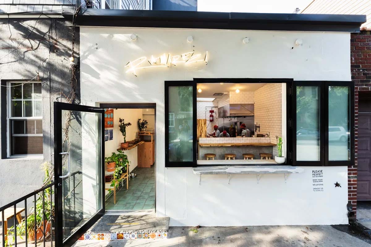white building with black-framed windows and an open door leading inside to a tiled room with bar stools.