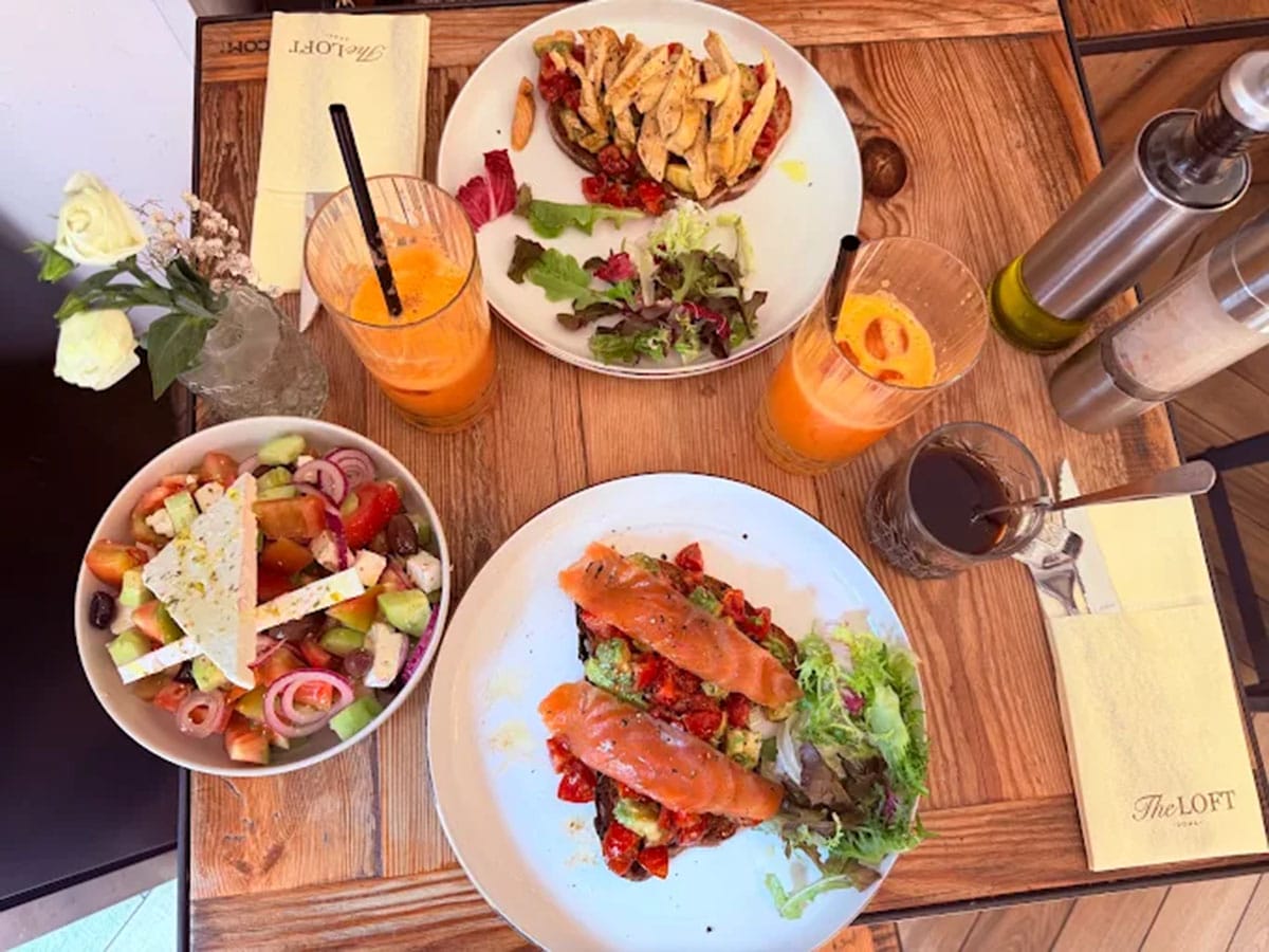 two white plates of fancy breakfast toasts with salad and drinks on a wooden table.
