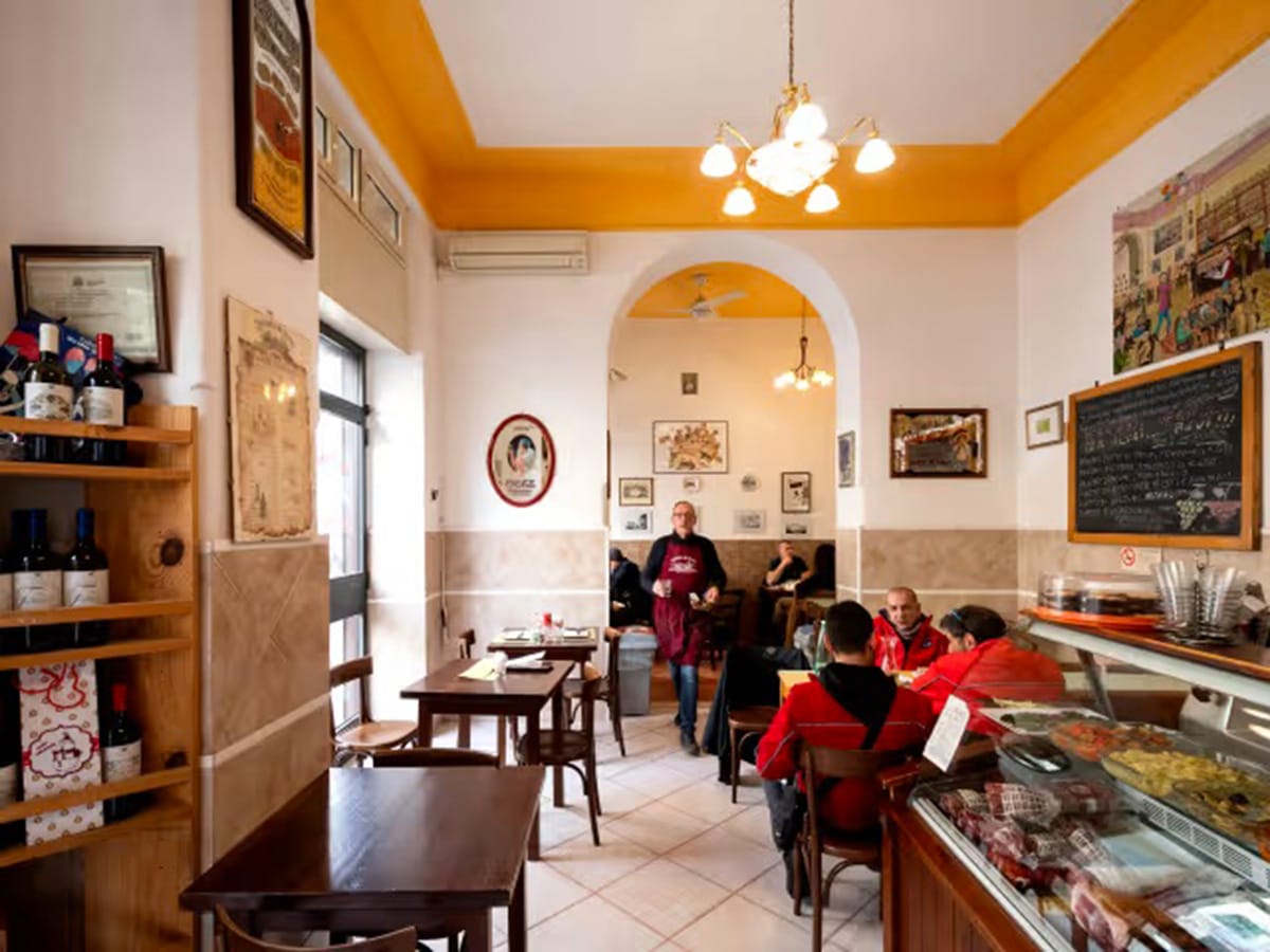 people sitting at tables in a sunlit restaurant with a case of pastries and a chalkboard on the wall.
