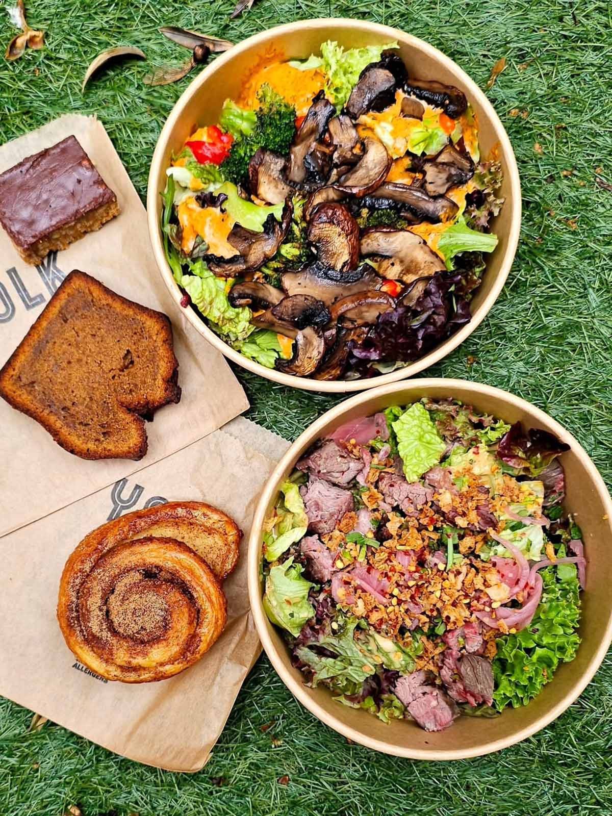 two disposable bowls filled with veggies with baked goods on napkins.