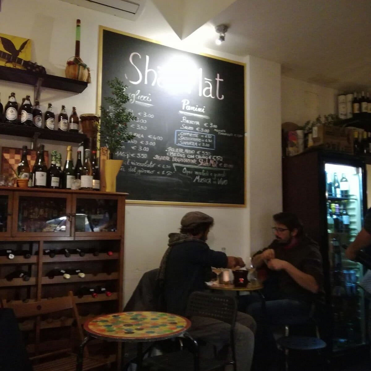 two men sitting at a round table with a shelf of wine bottles and a chalkboard on the wall.