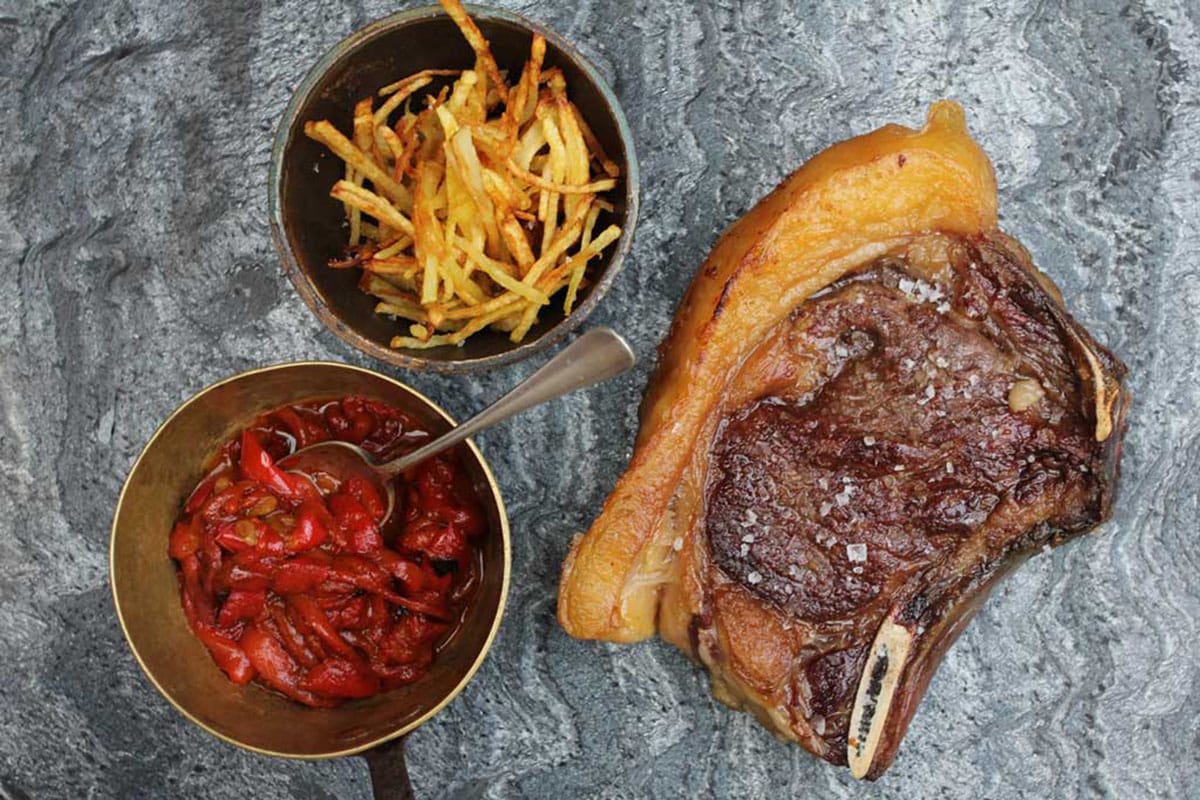piece of cooked steak with a bowl of red peppers and a bowl of thin fried potatoes.