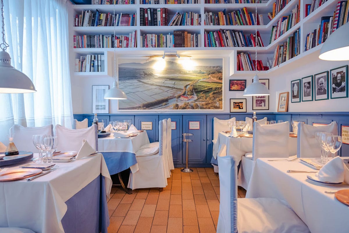 white slipcovered chairs at white-covered tables in a restaurant with bookshelves lining the walls.