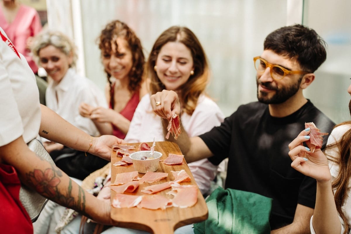 A tattoo-inked tour guide offers a wooden board piled with paper-thin prosciutto to a small group of seated guests.