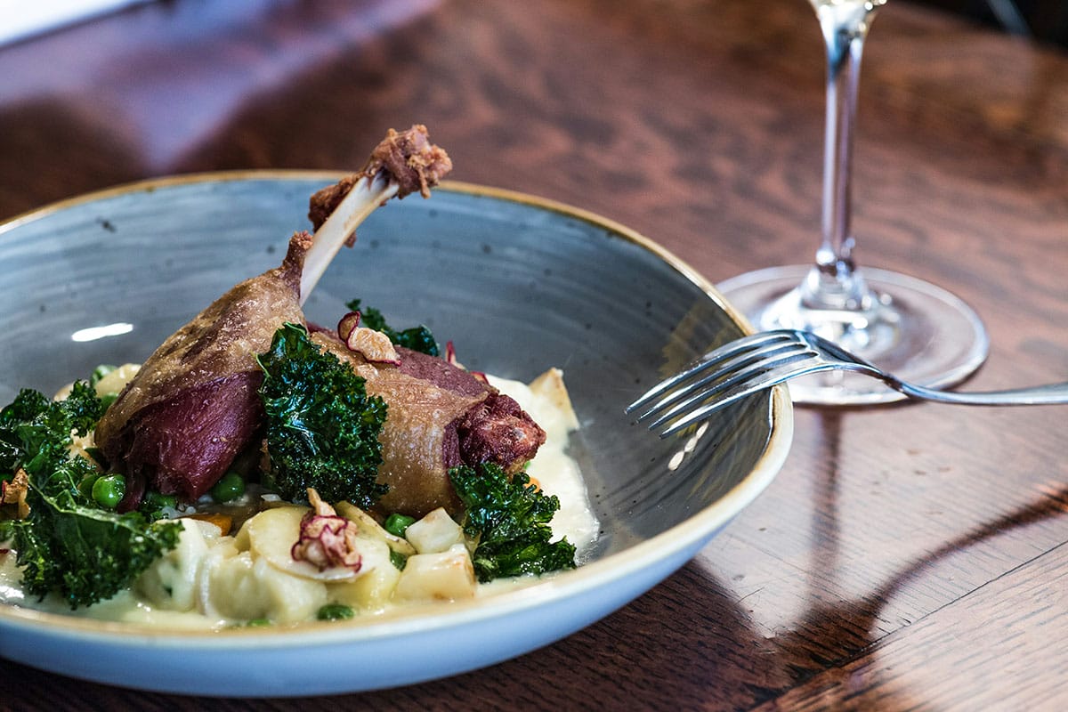 blue bowl of lamb with potatoes, kale, and cream sauce near a wine glass on a wooden table.