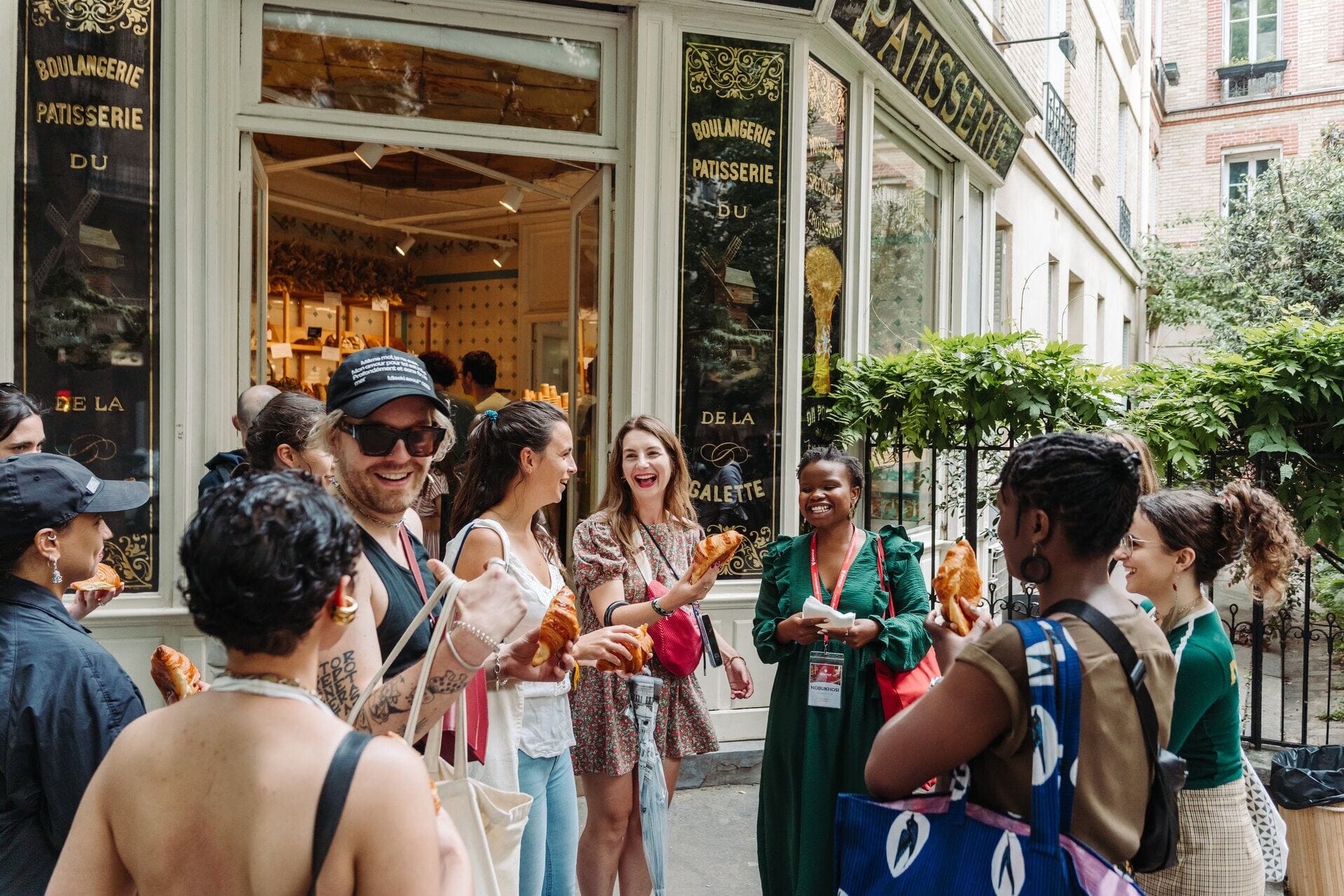 A cheerful group of people standing outside a French bakery, laughing and holding fresh croissants together during a food tour.