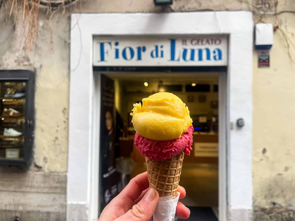 person holding a cone of yellow and pink gelato in front of the gelateria entrance.
