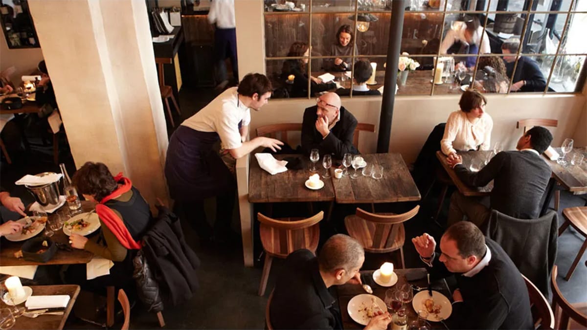 people eating and drinking at small wooden tables in a restaurant.