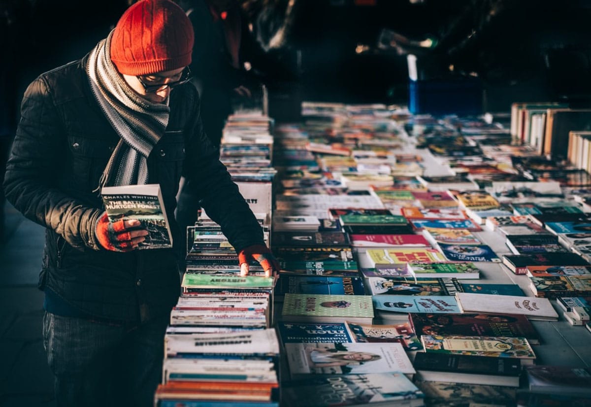 man in a coat, scarf, and hat looking at a table of used books outdoors.