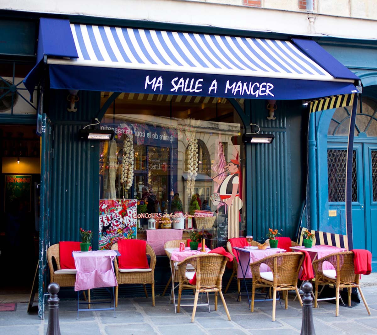 wicker chairs at red-covered tables in front of a restaurant with a blue awning.