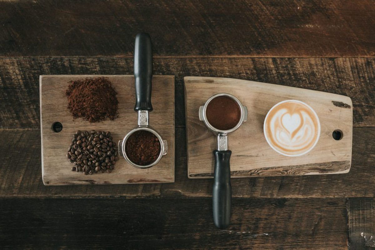 coffee beans and coffee ground and tamped into espresso machine attachments near a latte.