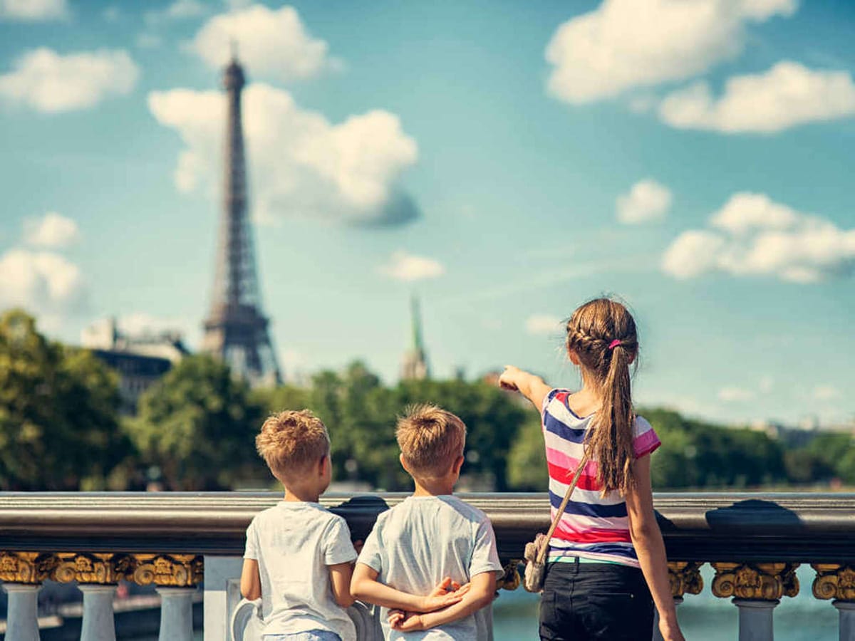 three children standing near a railing on a bridge with one girl pointing to the Eiffel Tower in the distance.