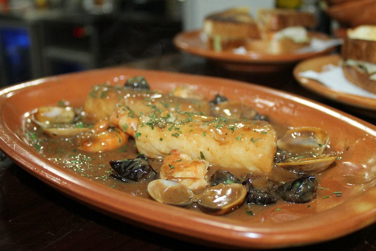 clay dish of cooked codfish with a sauce and clams.